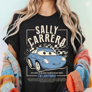 May include: Black t-shirt featuring a cartoon image of Sally Carrera, a blue Porsche 911, with the text "SALLY CARRERA" above the car. The shirt also includes the phrase "I'M LIGHTNING!"