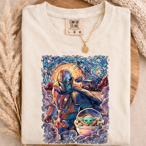 May include: Cream-coloured t-shirt with a colourful graphic of a Mandalorian character and Grogu in a starry night style. The shirt has a Comfort Colors tag and a gold necklace.