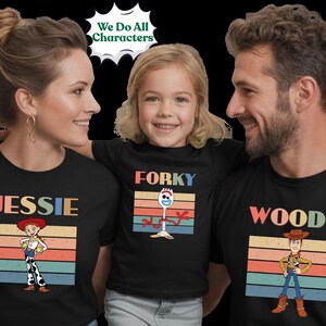 May include: Three black t-shirts featuring Toy Story character graphics. The left shirt shows Jessie with the text "JESSIE", the center shirt shows Forky with the text "FORKY", and the right shirt shows Woody with the text "WOOD". Each design includes a retro-style horizontal striped background.