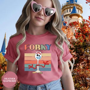 May include: A heather raspberry short-sleeve t-shirt featuring a graphic of the character Forky. The design includes the text FORKY above a retro-style sunset background with horizontal stripes in orange, yellow, teal, and blue colors.