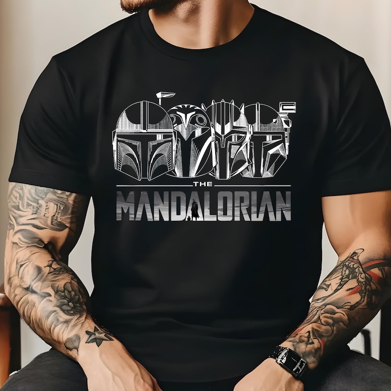 May include: Black t-shirt featuring a graphic of four Mandalorian helmets in white. The text "THE MANDALORIAN" is printed in silver below the helmets. The design is centered on the shirt.