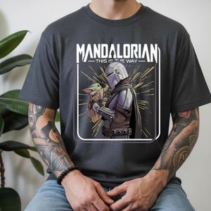May include: Dark gray t-shirt featuring a graphic of the Mandalorian holding Grogu, with the text "MANDALORIAN THIS IS THE WAY" in white. The design includes a silver helmet and a brown and tan Grogu.
