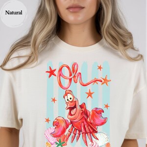Pu&ograve; includere: Una t-shirt color naturale con una stampa grafica di un granchio rosso con occhi grandi e bocca aperta. Il testo "Oh" &egrave; scritto sopra il granchio in un carattere rosso. Il design include strisce verticali azzurre, stelle marine arancioni e conchiglie.