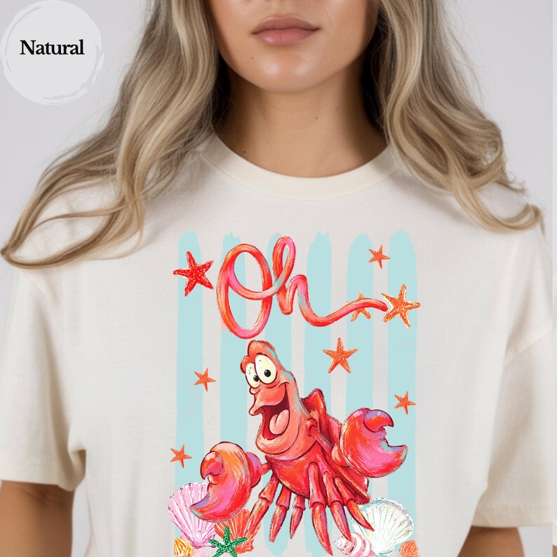 Pu&ograve; includere: Una t-shirt color naturale con una stampa grafica di un granchio rosso con occhi grandi e bocca aperta. Il testo "Oh" &egrave; scritto sopra il granchio in un carattere rosso. Il design include strisce verticali azzurre, stelle marine arancioni e conchiglie.