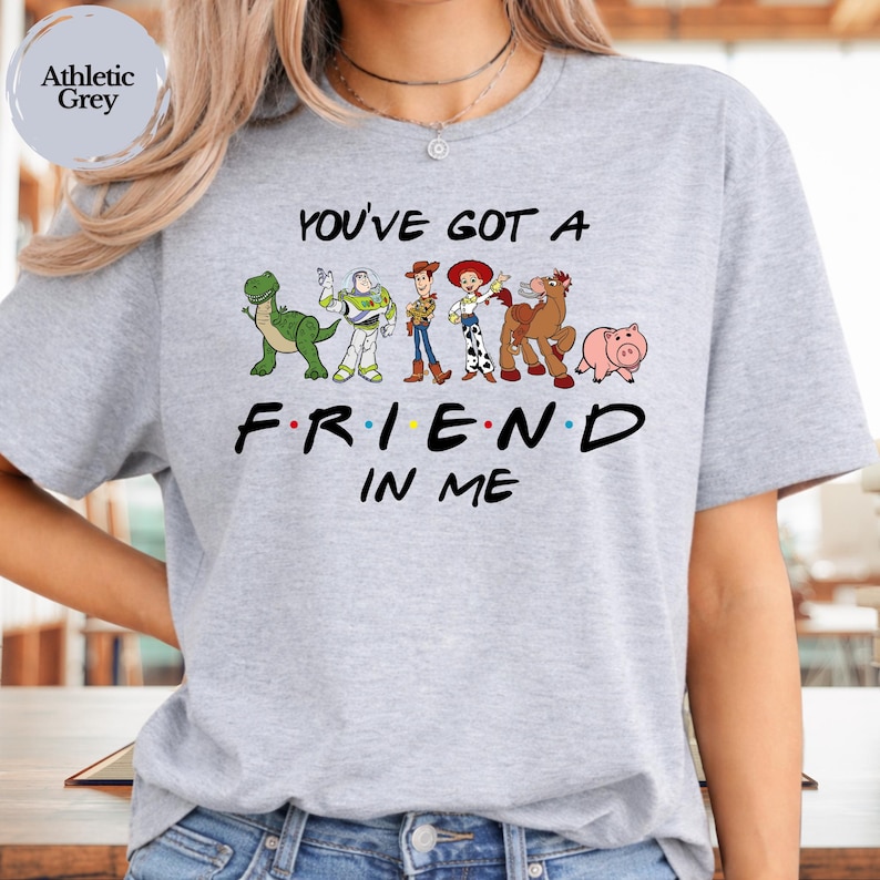May include: Athletic grey t-shirt featuring the phrase "You've Got A Friend In Me" with cartoon characters. The design includes a green dinosaur, Buzz Lightyear, Woody, Jessie, Bullseye, and Hamm the pig.