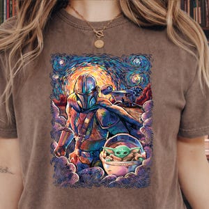 May include: Brown t-shirt with a vibrant graphic of The Mandalorian and Grogu, set against a starry night sky in the style of Van Gogh. The design includes the Mandalorian, Grogu in his pod, and a vehicle.