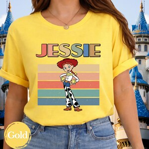 May include: A bright yellow short-sleeved t-shirt featuring a graphic of the character Jessie from Toy Story. The design includes the name JESSIE in colorful block letters above the character, set against a background of horizontal retro stripes.