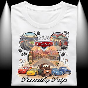 Puede incluir: Camiseta blanca con un gr&aacute;fico colorido de los personajes de Cars. El dise&ntilde;o incluye una pista de carreras, una gasolinera y el texto "2026 Cars Family Trip". El gr&aacute;fico muestra a Rayo McQueen, Mate y otros personajes.