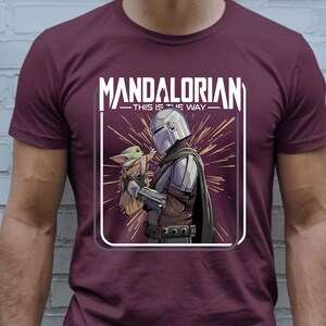 May include: Burgundy t-shirt featuring a graphic of The Mandalorian and Grogu. The image shows the Mandalorian in armor holding Grogu, with the text "MANDALORIAN - THIS IS THE WAY" in white.