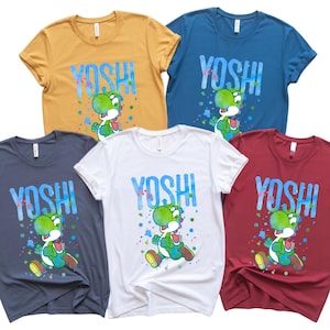 May include: Five short-sleeved t-shirts in gold, blue, grey, white, and burgundy. Each shirt displays the word "YOSHI" in blue and a graphic of the Yoshi character with colourful paint splatter accents.