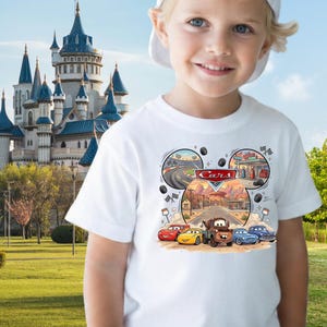 May include: White t-shirt featuring a colorful graphic of the Cars movie characters. The design includes Lightning McQueen, Mater, and other vehicles, with the word "Cars" in a red banner. The shirt is worn by a young child.