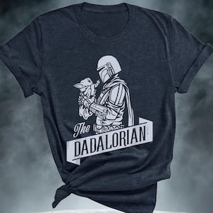 May include: Dark grey t-shirt with a white graphic of a Mandalorian figure cradling a small creature. The text "The DADALORIAN" is displayed on a banner. The shirt has rolled sleeves and a knotted hem.