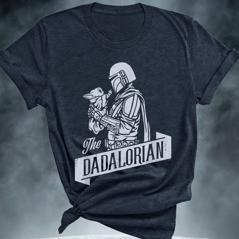May include: Dark grey t-shirt with a white graphic of a Mandalorian figure cradling a small creature. The text "The DADALORIAN" is displayed on a banner. The shirt has rolled sleeves and a knotted hem.