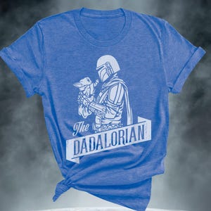 May include: Blue t-shirt featuring a white graphic of a character holding a small creature, with the text "The DADALORIAN" in a banner. The shirt has rolled sleeves and a knot at the hem.