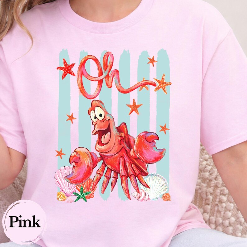 Pu&ograve; includere: Una t-shirt rosa chiaro con una stampa grafica di un granchio rosso dei cartoni animati con occhi grandi e bocca aperta. Il design include la parola Oh in corsivo rosso, strisce verticali azzurre, stelle marine arancioni e varie conchiglie.