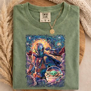 May include: Green Comfort Colors t-shirt with a colourful graphic of The Mandalorian and Grogu in a starry night style. The shirt has a crew neck and short sleeves. A gold necklace with a sun pendant is draped on the shirt.