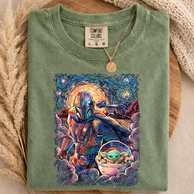 May include: Green Comfort Colors t-shirt with a colourful graphic of The Mandalorian and Grogu in a starry night style. The shirt has a crew neck and short sleeves. A gold necklace with a sun pendant is draped on the shirt.