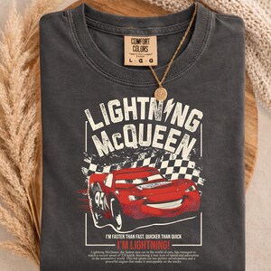 May include: Dark gray t-shirt featuring a graphic of Lightning McQueen, a red race car, with the text "LIGHTNING MCQUEEN" and "I'M LIGHTNING!". The shirt also has the Comfort Colors brand tag.
