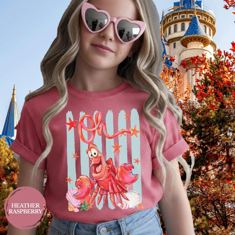 Pu&ograve; includere: Una t-shirt a maniche corte color lampone m&eacute;lange con una stampa grafica di un granchio rosso, pennellate verticali azzurre, stelle marine arancioni e conchiglie. Il testo "Oh" &egrave; stampato in un corsivo rosso sopra il personaggio.
