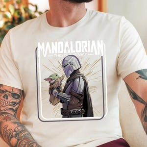 May include: Cream-colored t-shirt featuring a graphic of The Mandalorian and Grogu. The illustration shows the Mandalorian in armor holding Grogu. The text "MANDALORIAN - THIS IS THE WAY" is printed above the image.