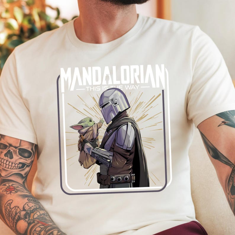 May include: Cream-colored t-shirt featuring a graphic of The Mandalorian and Grogu. The illustration shows the Mandalorian in armor holding Grogu. The text "MANDALORIAN - THIS IS THE WAY" is printed above the image.