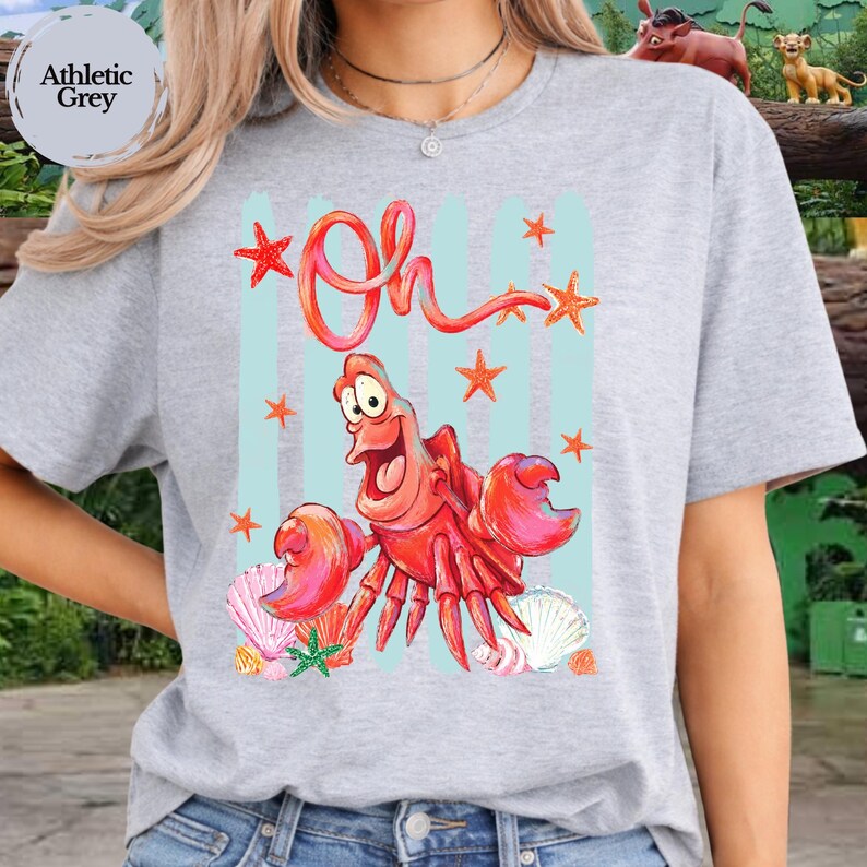 Pu&ograve; includere: Una t-shirt grigio atletico con una stampa grafica di un granchio rosso e la parola Oh scritta in corsivo rosso sopra. Il design include strisce verticali azzurre, stelle marine arancioni e varie conchiglie nei colori rosa, bianco e arancione.
