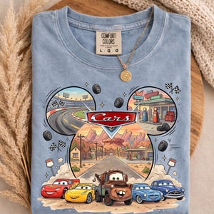 May include: Light blue t-shirt featuring a colorful graphic from the movie "Cars." The design includes the word "Cars" in a red and white logo, race tracks, and cartoon cars. The shirt has a Comfort Colors tag.