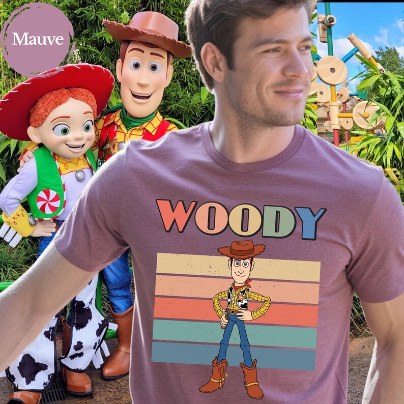 May include: A mauve short-sleeved t-shirt featuring a graphic print of the character Woody from Toy Story. The design includes the text WOODY in multi-colored letters above a retro-style striped background and a full-body illustration of the character.
