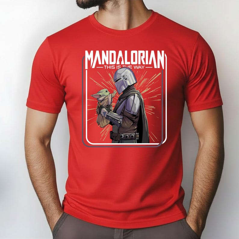 May include: Red t-shirt featuring a graphic of the Mandalorian holding Grogu, with the text "MANDALORIAN - THIS IS THE WAY" in a white rectangular frame. The design includes a red burst background. The shirt is short-sleeved.