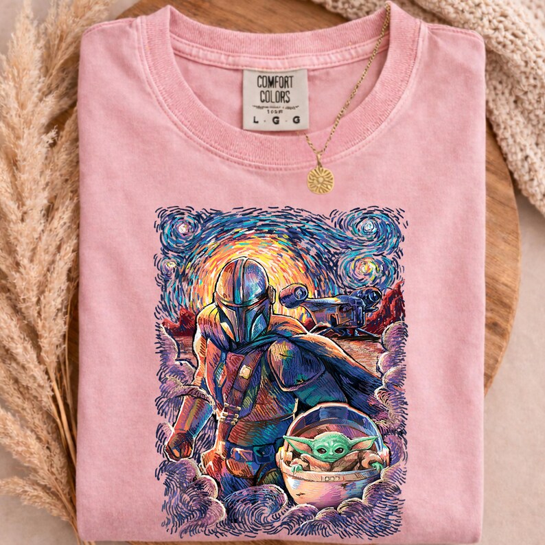 May include: Pale pink Comfort Colors t-shirt with a vibrant illustration of The Mandalorian and Grogu in a Starry Night style. The shirt has a crew neck and short sleeves. A gold necklace with a sun pendant is visible.