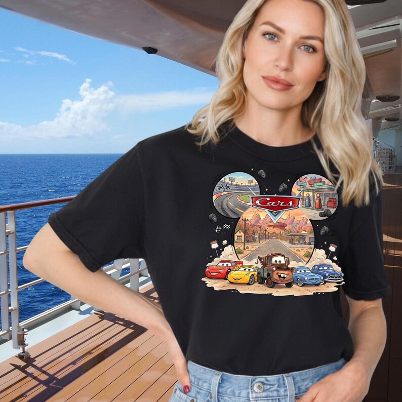 May include: Black t-shirt featuring a colorful graphic of characters from the movie "Cars". The design includes the word "Cars" in a red and white font, and images of race cars, a gas station, and a desert road.