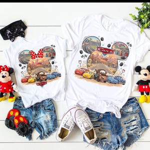 Pode incluir: Duas camisetas brancas com um gr&aacute;fico dos personagens do filme Carros dentro de uma silhueta de orelhas do Mickey Mouse. O design inclui uma paisagem des&eacute;rtica, pista de corrida e o logotipo Carros. Exibidas com shorts jeans e brinquedos.