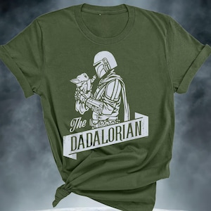 May include: Olive green t-shirt with a white graphic of a helmeted figure cradling a small creature. The text "The DADALORIAN" is in a white banner. The t-shirt is knotted at the bottom, with rolled-up sleeves.