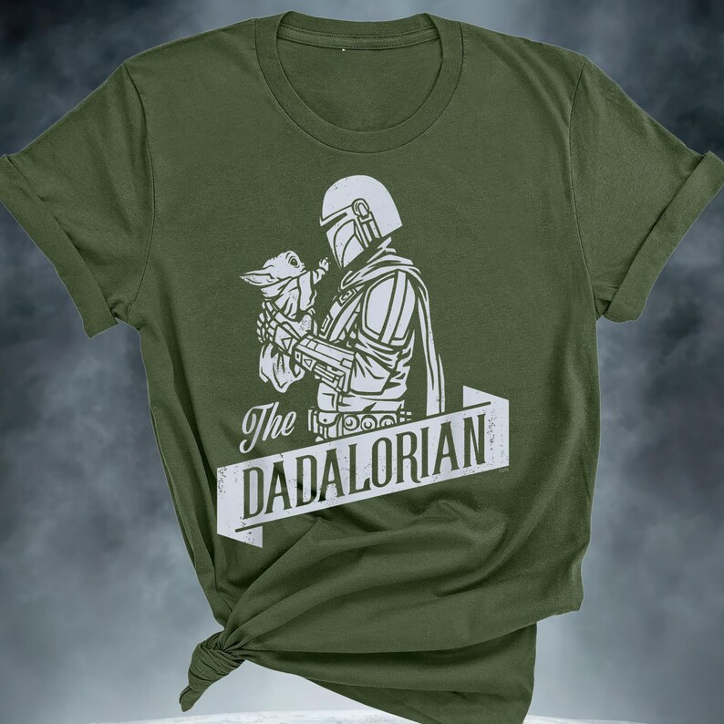 May include: Olive green t-shirt with a white graphic of a helmeted figure cradling a small creature. The text "The DADALORIAN" is in a white banner. The t-shirt is knotted at the bottom, with rolled-up sleeves.