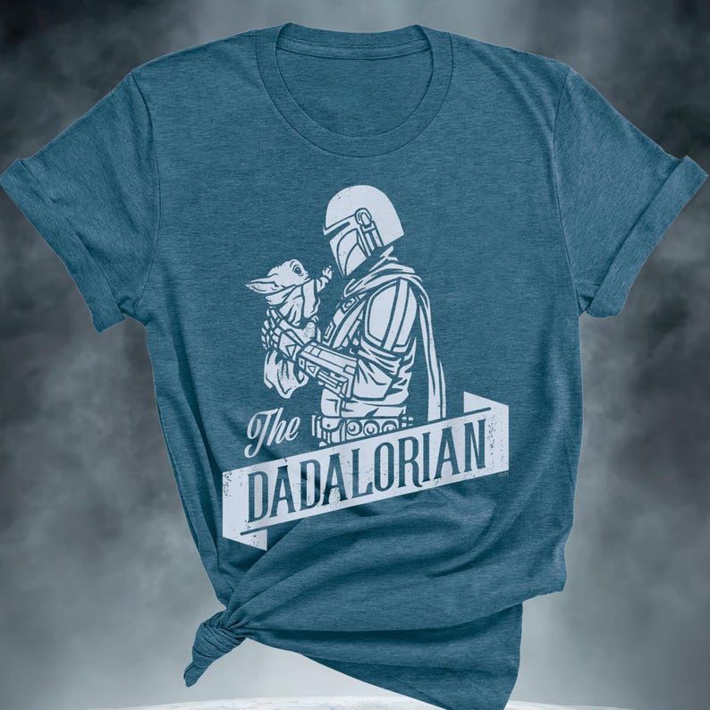 May include: Teal t-shirt with a white graphic of a character in armour holding a small creature. The text "The DADALORIAN" is displayed in a white banner below the graphic. The shirt is knotted at the bottom.