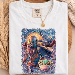 May include: White Comfort Colors t-shirt with a vibrant illustration of The Mandalorian and Grogu, set against a starry night sky. The design includes the text "COMFORT COLORS" and size details. A gold necklace with a sun pendant is draped over the shirt.