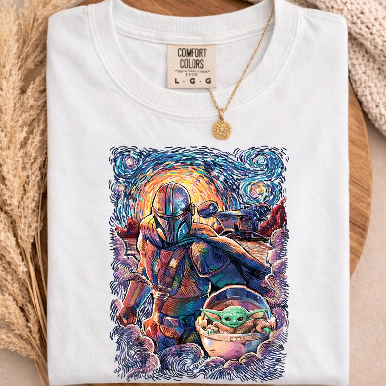 May include: White Comfort Colors t-shirt with a vibrant illustration of The Mandalorian and Grogu, set against a starry night sky. The design includes the text "COMFORT COLORS" and size details. A gold necklace with a sun pendant is draped over the shirt.
