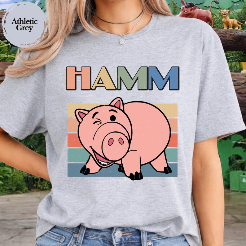 May include: An athletic grey short-sleeve t-shirt featuring a graphic of a pink pig character. Above the pig, the text HAMM is printed in bold, multi-colored block letters. The design includes a retro-style horizontal striped background.