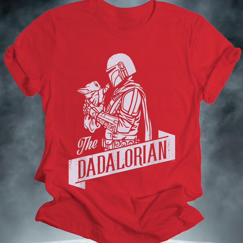 May include: Red t-shirt with a white graphic of a Mandalorian character holding a small creature. The text "The DADALORIAN" is printed below the image in a banner style. The shirt has rolled-up sleeves.