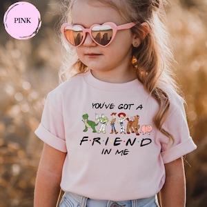 May include: A light pink t-shirt featuring the phrase "YOU'VE GOT A FRIEND IN ME" with cartoon character graphics. The shirt is worn by a child wearing pink heart-shaped sunglasses.