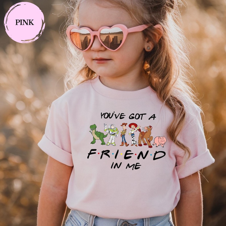 May include: A light pink t-shirt featuring the phrase "YOU'VE GOT A FRIEND IN ME" with cartoon character graphics. The shirt is worn by a child wearing pink heart-shaped sunglasses.
