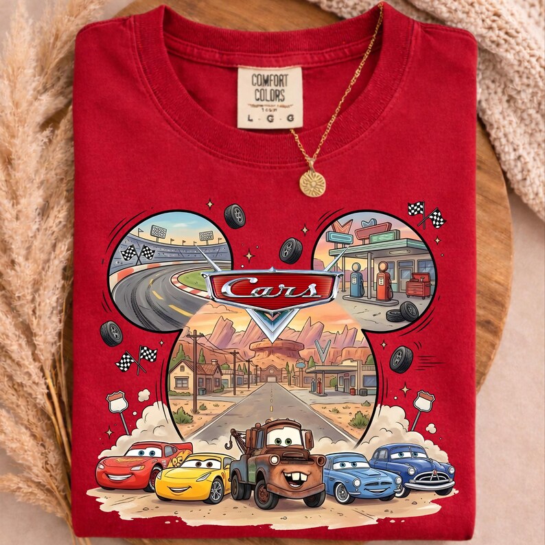 May include: Red t-shirt featuring a colorful graphic of characters and scenes from the animated movie "Cars." The design includes race cars, a desert road, and the "Cars" logo. The shirt has a crew neck and short sleeves.