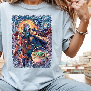 May include: Pale blue t-shirt with a vibrant, painterly illustration of a Mandalorian character and Grogu, set against a swirling night sky. The artwork uses blues, oranges, and greens. The design includes a spaceship and a baby character in a pod.