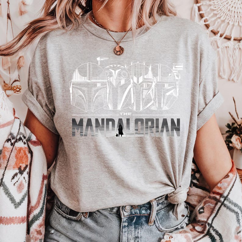 May include: A light gray t-shirt featuring a graphic design of Mandalorian helmets and the text "THE MANDALORIAN". The design is in white and gray tones, with a small silhouette of a figure at the bottom.