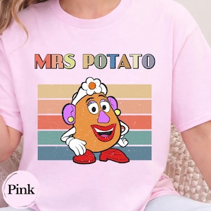 May include: A light pink t-shirt featuring a graphic of Mrs. Potato Head with a white flower on her head, purple ears, and red shoes. Behind the character are horizontal stripes in muted retro colors. The text MRS POTATO is printed above.