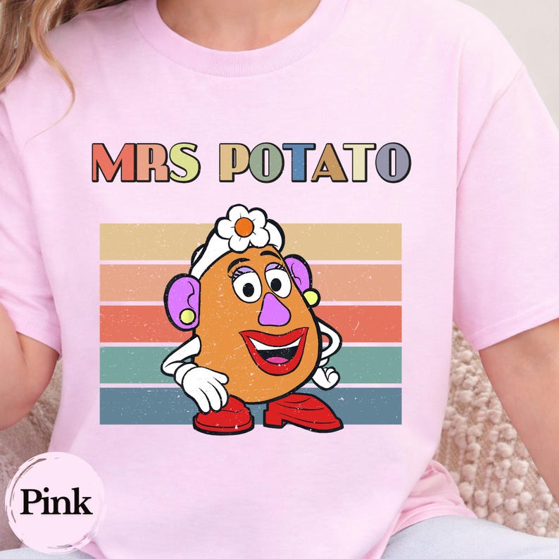 May include: A light pink t-shirt featuring a graphic of Mrs. Potato Head with a white flower on her head, purple ears, and red shoes. Behind the character are horizontal stripes in muted retro colors. The text MRS POTATO is printed above.
