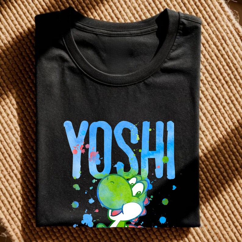 May include: Black t-shirt with the word "YOSHI" in blue, and a green and white cartoon character. The design includes colourful paint splatters. The shirt is folded on a textured, tan surface.