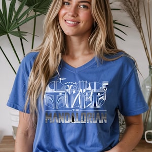 May include: A blue V-neck t-shirt featuring a white graphic design of Mandalorian helmets and the text "THE MANDALORIAN". The design is centered on the chest. The shirt is made of a soft material.