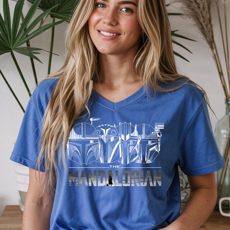 May include: A blue V-neck t-shirt featuring a white graphic design of Mandalorian helmets and the text "THE MANDALORIAN". The design is centered on the chest. The shirt is made of a soft material.