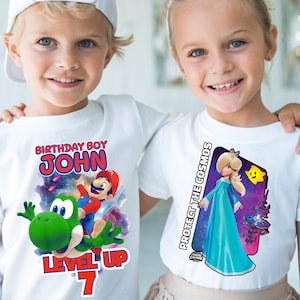 May include: Two children wearing white t-shirts with colorful graphic designs. One shirt says "BIRTHDAY BOY JOHN" with Mario and Yoshi. The other shirt says "PROTECT THE COSMOS" with a princess character. Both shirts have a "Mario Galaxy" logo.